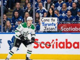 Jason Robertson of the Dallas Stars in his white away uniform skates on the ice during an NHL game against the Toronto Maple Leafs, with fans visible in the stands behind the glass.