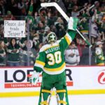 Minnesota Wild goaltender Jesper Wallstedt (30) stands on the ice at Xcel Energy Center, raising his stick to the crowd. Behind the glass boards, a fan holds a handmade sign in red and green ink that reads "Please don't trade Jesper!" amidst cheering spectators.