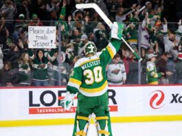 Minnesota Wild goaltender Jesper Wallstedt (30) stands on the ice at Xcel Energy Center, raising his stick to the crowd. Behind the glass boards, a fan holds a handmade sign in red and green ink that reads "Please don't trade Jesper!" amidst cheering spectators.