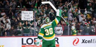 Minnesota Wild goaltender Jesper Wallstedt (30) stands on the ice at Xcel Energy Center, raising his stick to the crowd. Behind the glass boards, a fan holds a handmade sign in red and green ink that reads "Please don't trade Jesper!" amidst cheering spectators.