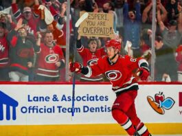 Is This The End? Why The Carolina Hurricanes Are Finally Trading Jesperi Kotkaniemi A photo of Carolina Hurricanes player Jesperi Kotkaniemi celebrating on the ice, raising his stick in the air. Behind the arena glass, amidst cheering spectators, a fan holds up a handwritten cardboard sign that reads, "JESPERI, YOU ARE OVERPAID!".
