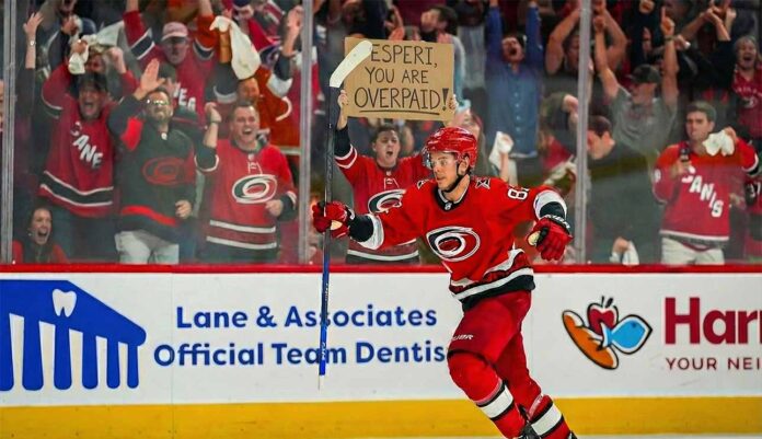 A photo of Carolina Hurricanes player Jesperi Kotkaniemi celebrating on the ice, raising his stick in the air. Behind the arena glass, amidst cheering spectators, a fan holds up a handwritten cardboard sign that reads, "JESPERI, YOU ARE OVERPAID!".