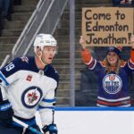 Why a Jonathan Toews Gamble Could Save the Oilers’ Third Line : Winnipeg Jets forward Jonathan Toews, wearing his white away #19 jersey, looks on from the ice. Behind the glass in the stands, a smiling female fan wearing an Edmonton Oilers jersey and toque holds up a handwritten cardboard sign that reads, "Come to Edmonton Jonathan!".