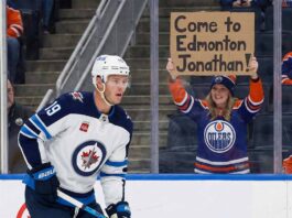 : Winnipeg Jets forward Jonathan Toews, wearing his white away #19 jersey, looks on from the ice. Behind the glass in the stands, a smiling female fan wearing an Edmonton Oilers jersey and toque holds up a handwritten cardboard sign that reads, "Come to Edmonton Jonathan!".