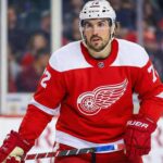 A waist-up photograph of a hockey player wearing a Detroit Red Wings red home jersey with number 72 and the winged wheel logo. He is standing on the ice, holding his stick, and looking intently forward against a backdrop of blurry fans.
