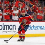 Florida Panthers defenseman Justin Faulk, wearing number 72 in the red home uniform, skates with the puck down the ice. Fans wearing Panthers jerseys and waving flags cheer from behind the glass near Holman and Ticketmaster dasher boards.