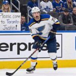 St. Louis Blues defenseman Justin Faulk in a white away jersey skates on the ice near the boards, looking towards the stands where a fan wearing Toronto Maple Leafs gear holds a handwritten sign that reads, "Please come to Toronto Justin!".