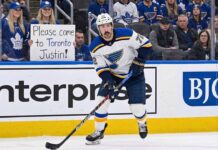 St. Louis Blues defenseman Justin Faulk in a white away jersey skates on the ice near the boards, looking towards the stands where a fan wearing Toronto Maple Leafs gear holds a handwritten sign that reads, "Please come to Toronto Justin!".