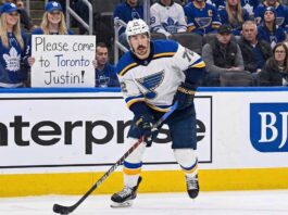 Is Doug Armstrong Dreaming? The Shocking Price for Justin Faulk St. Louis Blues defenseman Justin Faulk in a white away jersey skates on the ice near the boards, looking towards the stands where a fan wearing Toronto Maple Leafs gear holds a handwritten sign that reads, "Please come to Toronto Justin!".