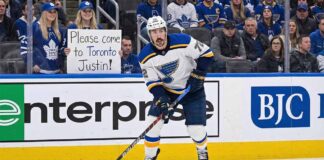 St. Louis Blues defenseman Justin Faulk in a white away jersey skates on the ice near the boards, looking towards the stands where a fan wearing Toronto Maple Leafs gear holds a handwritten sign that reads, "Please come to Toronto Justin!".