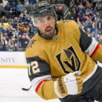 Vegas Golden Knights defenseman Justin Faulk, wearing the number 72 gold home jersey, skates on the ice during a game, looking intently towards the left. Fans and a St. Louis Blues player are visible in the background.
