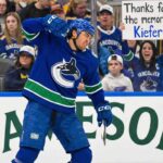 Kiefer Sherwood #44 of the Vancouver Canucks skates with the puck in a white away uniform. In the background behind the glass, a female fan holds up a handwritten sign reading "Thanks for the memories Kiefer!".