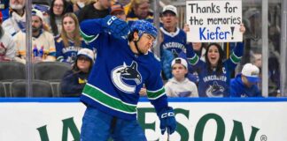 Kiefer Sherwood #44 of the Vancouver Canucks skates with the puck in a white away uniform. In the background behind the glass, a female fan holds up a handwritten sign reading "Thanks for the memories Kiefer!".