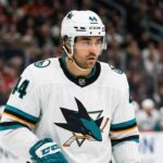 Concept image of Vancouver Canucks forward Kiefer Sherwood celebrating fiercely on the ice while wearing a teal San Jose Sharks away jersey.