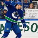 Vancouver Canucks forward Kiefer Sherwood celebrates a goal on the ice, with a female fan behind the glass holding a sign that reads "Please stay in Vancouver Kiefer!". The Jameson ad is visible on the boards.