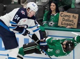 Why the Winnipeg Jets Must Trade Logan Stanley Now A smiling female Dallas Stars fan in the stands holds a cardboard sign reading "Come to Dallas Logan!" while Winnipeg Jets defenseman Logan Stanley checks a Stars player into the boards just in front of her behind the glass.