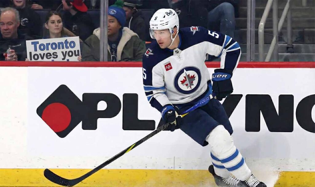 Winnipeg Jets defenseman Luke Schenn, wearing the number 5 away jersey, skates along the boards during a game. In the background behind the glass, a smiling female fan wearing a blue Toronto Maple Leafs jersey holds up a handwritten sign that reads, "TORONTO LOVES YOU LUKE!".