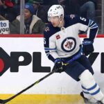 Winnipeg Jets defenseman Luke Schenn, wearing the number 5 away jersey, skates along the boards during a game. In the background behind the glass, a smiling female fan wearing a blue Toronto Maple Leafs jersey holds up a handwritten sign that reads, "TORONTO LOVES YOU LUKE!".