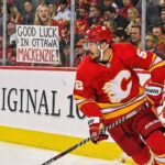 Calgary Flames defenseman MacKenzie Weegar skating with the puck along the boards during a game. Behind the glass, a smiling blonde fan holds up a handwritten white sign that reads, "GOOD LUCK IN OTTAWA MACKENZIE!", referencing trade rumors connecting him to the Ottawa Senators.
