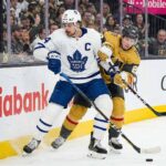 Toronto Maple Leafs captain Auston Matthews in a white away jersey battles for a loose puck along the arena boards against Mitch Marner wearing a gold home jersey during an NHL game.
