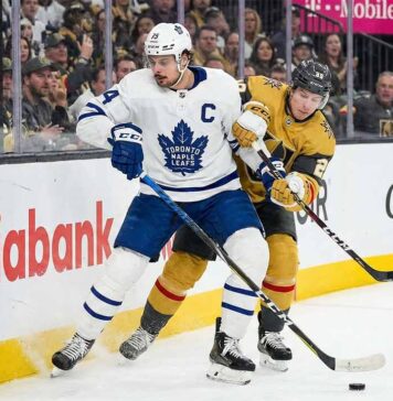 Toronto Maple Leafs captain Auston Matthews in a white away jersey battles for a loose puck along the arena boards against Mitch Marner wearing a gold home jersey during an NHL game.