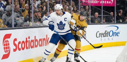 Toronto Maple Leafs captain Auston Matthews in a white away jersey battles for a loose puck along the arena boards against Mitch Marner wearing a gold home jersey during an NHL game.