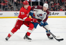 Detroit Red Wings defenseman Moritz Seider (53) in a red home jersey and Colorado Avalanche forward Nathan MacKinnon (29) in a white and maroon away jersey battle fiercely for the puck along the boards during an NHL game at Little Caesars Arena.