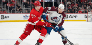 Detroit Red Wings defenseman Moritz Seider (53) in a red home jersey and Colorado Avalanche forward Nathan MacKinnon (29) in a white and maroon away jersey battle fiercely for the puck along the boards during an NHL game at Little Caesars Arena.