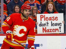 Calgary Flames center Nazem Kadri, wearing his red home jersey, looks towards the action on the ice. In the stands behind the glass, a young female fan with red hair holds up a handwritten sign with purple and orange letters that reads, "Please don't leave Nazem!".