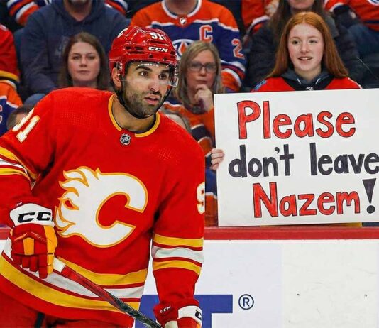 Calgary Flames center Nazem Kadri, wearing his red home jersey, looks towards the action on the ice. In the stands behind the glass, a young female fan with red hair holds up a handwritten sign with purple and orange letters that reads, "Please don't leave Nazem!".