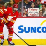 Calgary Flames center Nazem Kadri #91 looks up while on the ice. Behind the glass partition, a smiling female fan holds up a white handmade sign with red text that reads, "Calgary loves you Nazem!".