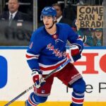 New York Rangers defenseman Braden Schneider (#4) skates on the ice during a game, while a female fan wearing San Jose Sharks apparel behind the glass holds up a handmade cardboard sign that reads, "COME TO SAN JOSE BRADEN!".