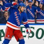 New York Rangers player Kiefer Sherwood, wearing number 44, pumps his fist and yells in celebration on the ice during a game. Fans wearing Rangers jerseys cheer behind the glass, and a Jameson Irish Whiskey advertisement is visible on the boards.