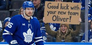 Toronto Maple Leafs defenseman Oliver Ekman-Larsson (95) is on the ice during a game, while in the background, a smiling female fan holds a cardboard sign that reads, "Good luck with your new team Oliver!".