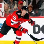 New Jersey Devils forward Ondrej Palat (#18) skates with the puck during an NHL game. Behind the glass in the stands, a blonde female fan holds up a sign with red and black letters that reads, "We will miss you Ondrej!"