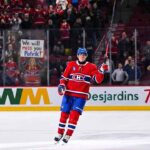 Montreal Canadiens forward Patrik Laine skates on the ice, saluting the crowd with his stick raised. Behind the rink glass, a female fan in a Habs jersey holds a handmade sign reading "We will miss you Patrik!".