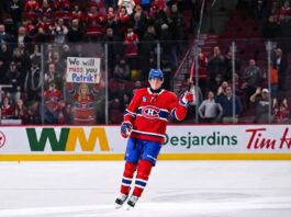 Montreal Canadiens forward Patrik Laine skates on the ice, saluting the crowd with his stick raised. Behind the rink glass, a female fan in a Habs jersey holds a handmade sign reading "We will miss you Patrik!".
