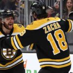 Boston Bruins forward Pavel Zacha (18), with his back to the camera, is embraced by his teammate David Pastrnak (88), who is smiling broadly, as they celebrate a goal near the glass during a game. Cheering fans are visible in the background.