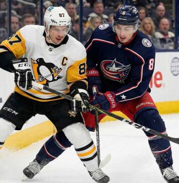 Pittsburgh Penguins captain Sidney Crosby (#87) in a white away jersey and Columbus Blue Jackets defenseman Zach Werenski (#8) in a navy home jersey clash sticks while battling for possession of the puck along the ice boards, with FanDuel Sportsbook and Navy Federal Credit Union advertisements visible in the background.