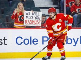 Calgary Flames defenseman Rasmus Andersson (#4) skates on the ice in his red home uniform. Behind the glass in the stands, a female fan holds up a handmade white sign with red and black text that reads, "WE WILL MISS YOU RASMUS!" while waving.