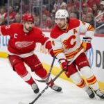 Calgary Flames defenseman Rasmus Andersson (#4) in a white away jersey skates with the puck along the boards, closely pursued by Detroit Red Wings captain Dylan Larkin (#71) in a red home jersey during an NHL game.