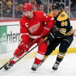 Detroit Red Wings captain Dylan Larkin (71) in a red jersey and Boston Bruins forward David Pastrnak (88) in a black jersey battle for the puck along the boards during an NHL game.