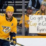 The Heartbreaking Decision Facing the Nashville Predators and Ryan O’Reilly Nashville Predators alternate captain Ryan O'Reilly (90) stands on the ice looking forward. Behind the glass barrier, a smiling female fan in a Predators jersey and beanie holds up a white poster sign with handwritten text that reads, "Go get your Cup in Colorado Ryan!".