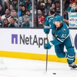 San Jose Sharks forward Jeff Skinner (#53) skates with the puck during a game. Behind him in the stands, a smiling female fan wearing a Sharks beanie holds a handwritten sign that reads, 'Good luck with your next team Jeff!'.