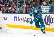 San Jose Sharks forward Jeff Skinner (#53) skates with the puck during a game. Behind him in the stands, a smiling female fan wearing a Sharks beanie holds a handwritten sign that reads, 'Good luck with your next team Jeff!'.