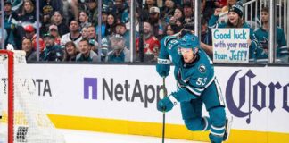 San Jose Sharks forward Jeff Skinner (#53) skates with the puck during a game. Behind him in the stands, a smiling female fan wearing a Sharks beanie holds a handwritten sign that reads, 'Good luck with your next team Jeff!'.