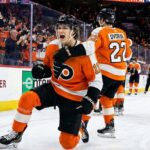 Philadelphia Flyers forward Trevor Zegras (#11) yells in celebration on one knee after scoring a goal at Wells Fargo Center, emphasizing his on-ice value amidst contract extension rumors. Teammate Christian Dvorak (#22) is behind him joining the celebration.