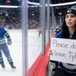 A female Vancouver Canucks fan with raven-black hair, wearing a team jersey and toque, holds a handmade sign against the arena glass that reads "Please don't leave Tyler!". In the background, on the ice, Canucks defenseman Tyler Myers, wearing jersey number 57, is skating away.