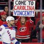 Is Vincent Trocheck Heading Home to the Hurricanes? Vincent Trocheck, wearing a New York Rangers jersey, skates on the ice during a game, while a female fan in a Carolina Hurricanes jersey behind the glass holds a sign that reads, "COME TO CAROLINA VINCENT!".