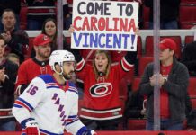 Vincent Trocheck, wearing a New York Rangers jersey, skates on the ice during a game, while a female fan in a Carolina Hurricanes jersey behind the glass holds a sign that reads, "COME TO CAROLINA VINCENT!".